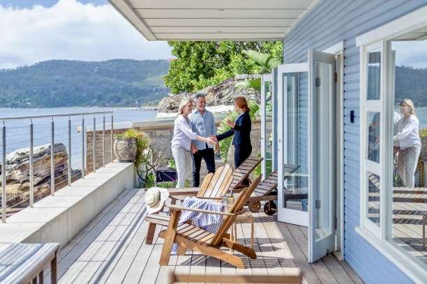 Real estate agent showing a mature couple a new house. The house is contemporary. All are happy and smiling and shaking hands. The couple are casually dressed and the agent is in a suit. Waterfront can be seen in the background